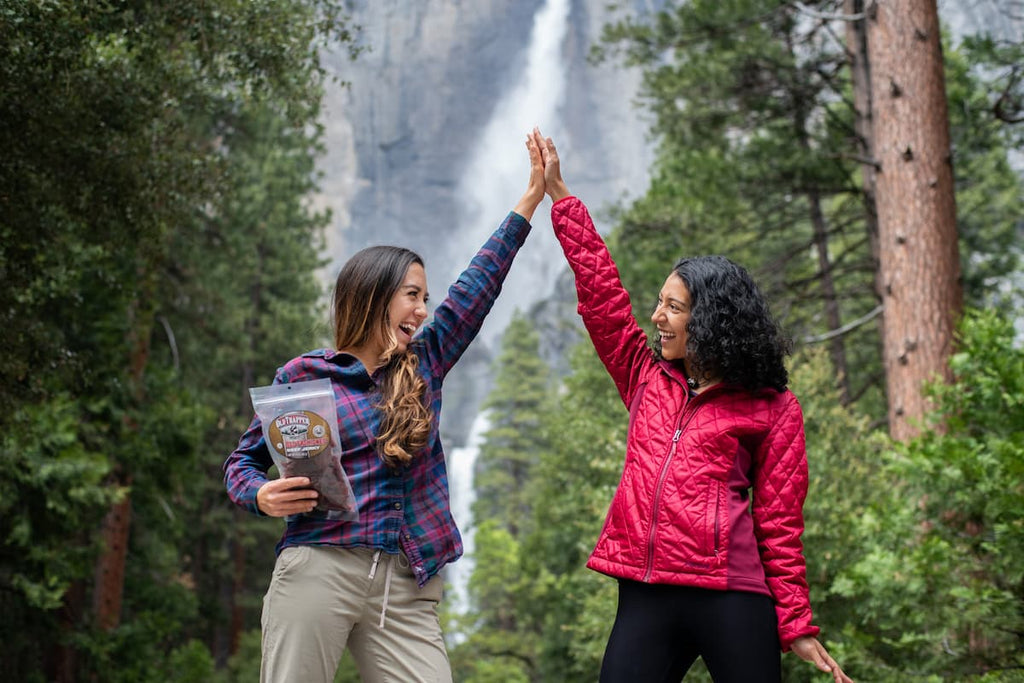 Two women share a high-five in front of a forest waterfall after discovering that Old Trapper is the best dry beef jerky for camping