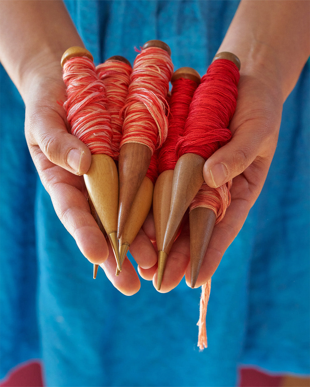 Hands of Ema Shine holding wooden spindles wound with red and variegated yarn, symbolizing the traditional craftsmanship and vibrant creativity in women's textile work.