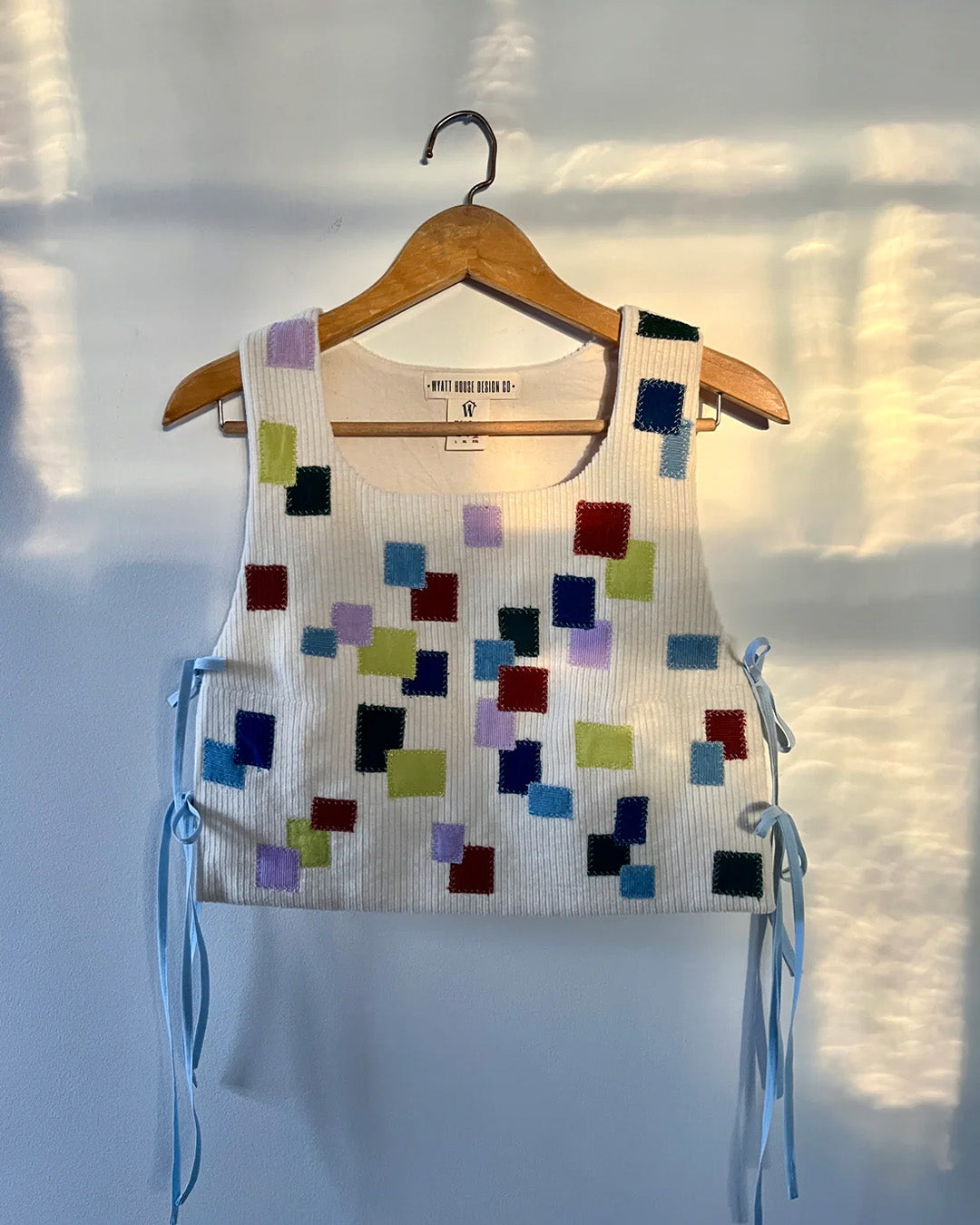 A handwoven top with colorful square patterns displayed on a hanger, bathed in soft light that casts a serene glow, illustrating the seamless blend of vibrant colors and imaginative design in textile art.
