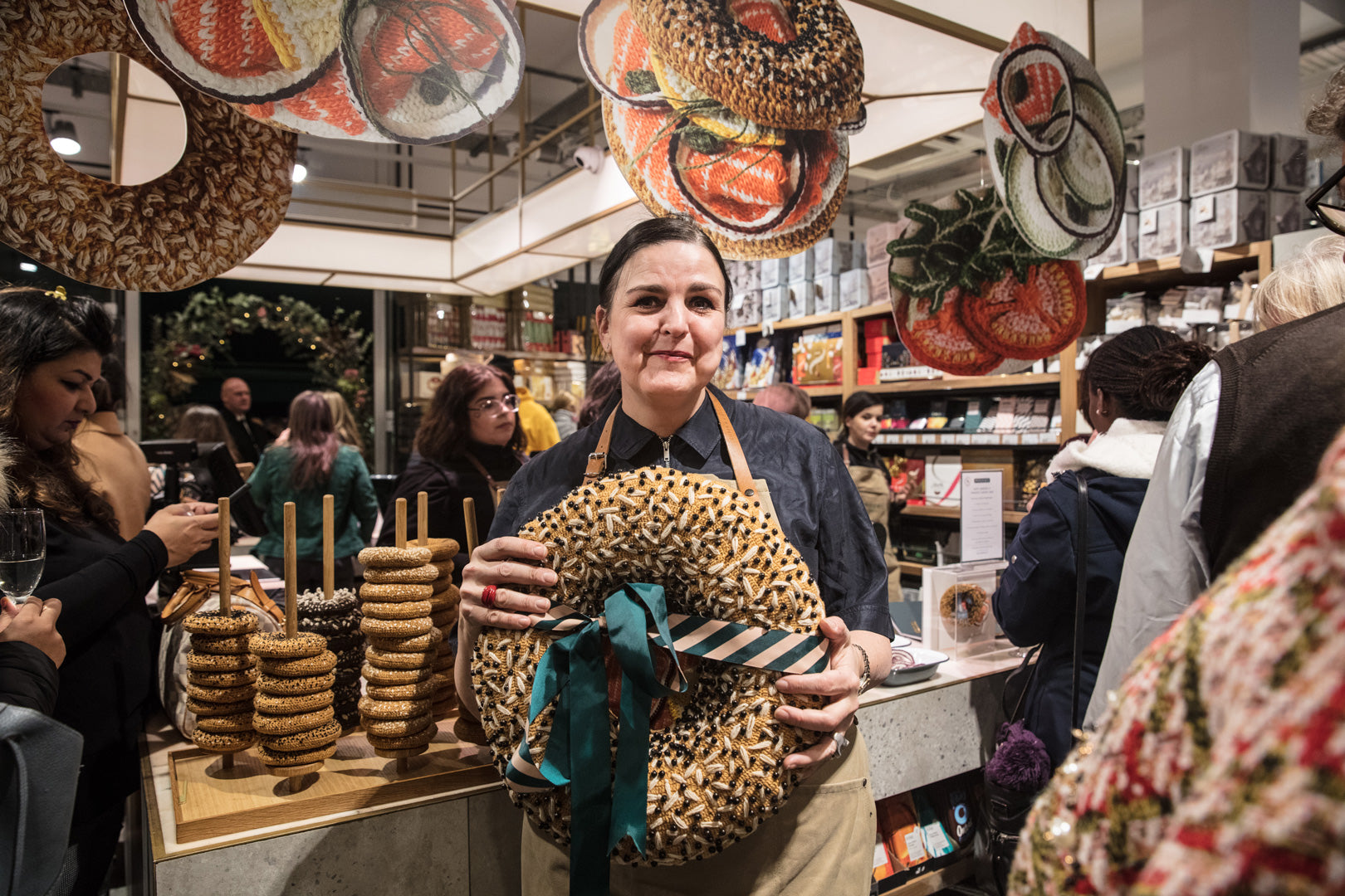 An artist: Kate Jenkins, proudly presents her handcrafted wreath that looks like pretzels at an exhibition, with large knit fruit decorations overhead, showcasing a festive and creative atmosphere.