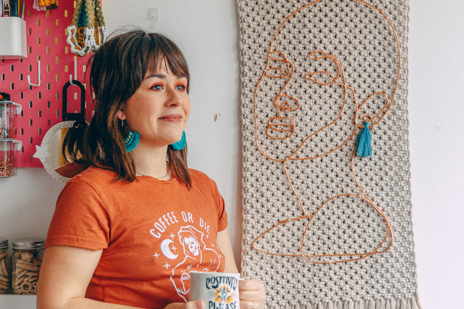 A creative female artist: Naomi Burke, with blue tassel earrings and an orange t-shirt sips from a mug, with a whimsical copper wire face sculpture on woven textile behind her, in a studio that bursts with personality and art.