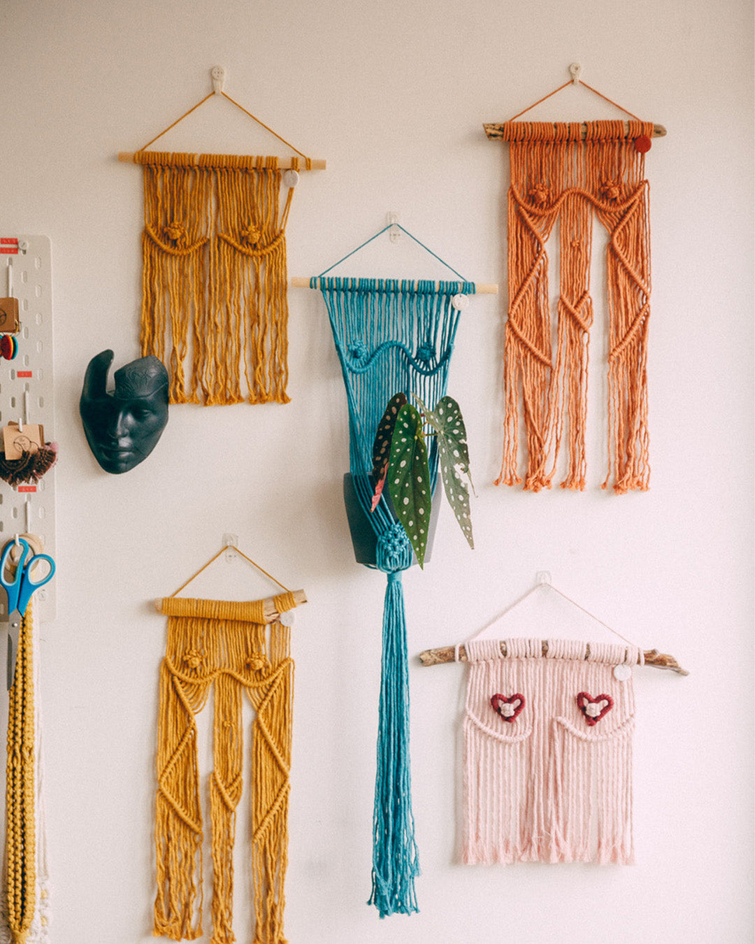 A collection of yarn wall hangings in warm hues displayed on a white wall, featuring playful patterns and varying textures that exhibit Naomi's creative use of knit and macramé techniques.