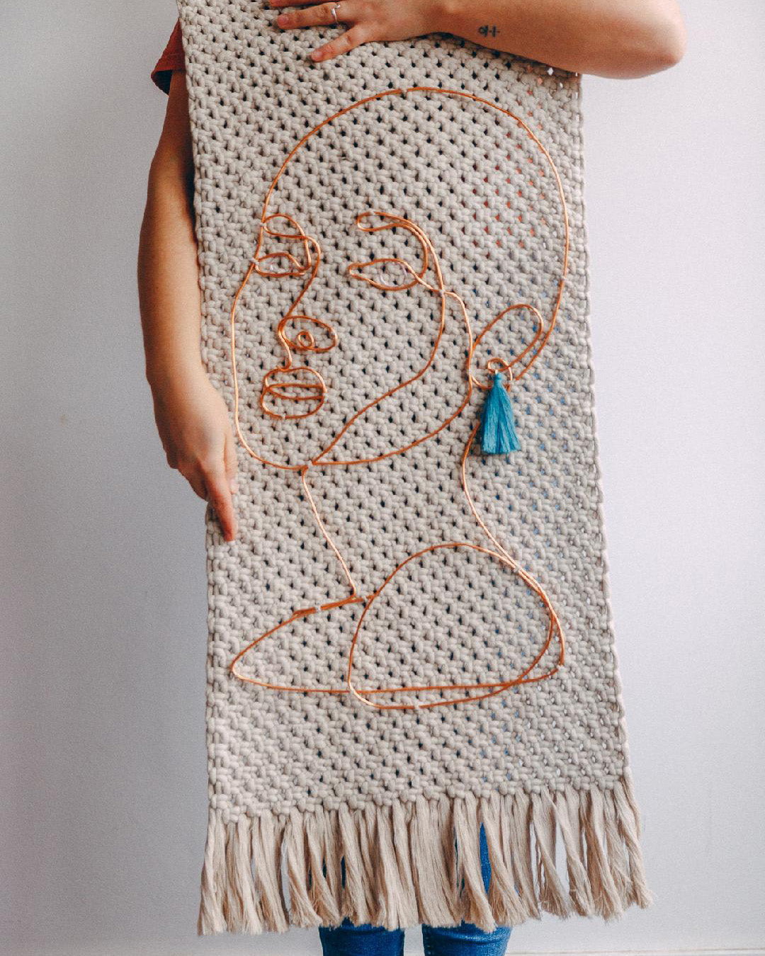 holds up a handwoven wall hanging with an abstract copper wire face and a blue tassel, symbolizing the fusion of minimalist art and tactile weaving in a display of creative expression.