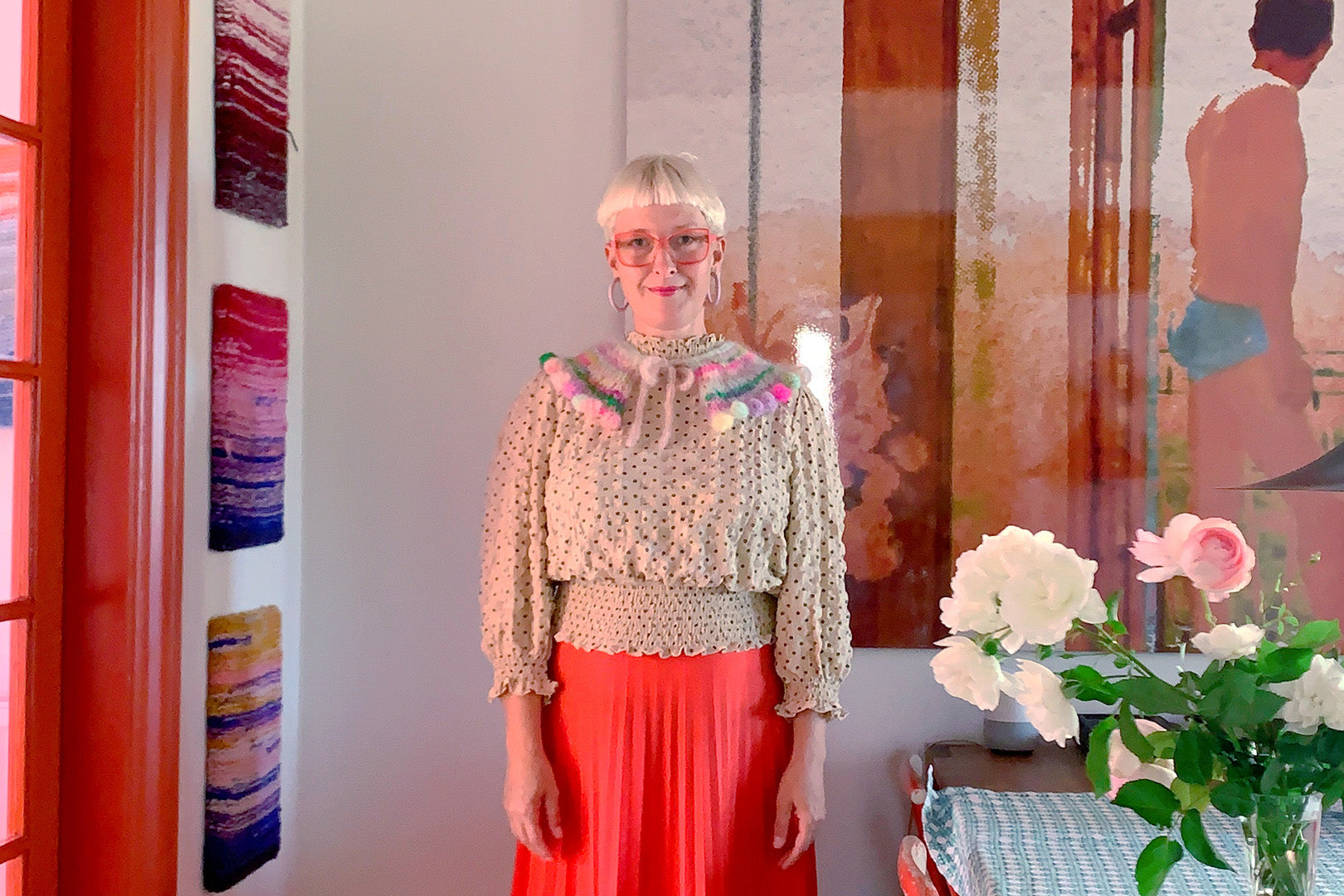 Artist Sofie wearing pink-fringed knitwear and glasses, smiles beside a wall with vibrant textile art pieces, reflecting a bright, inspiring creative studio space.