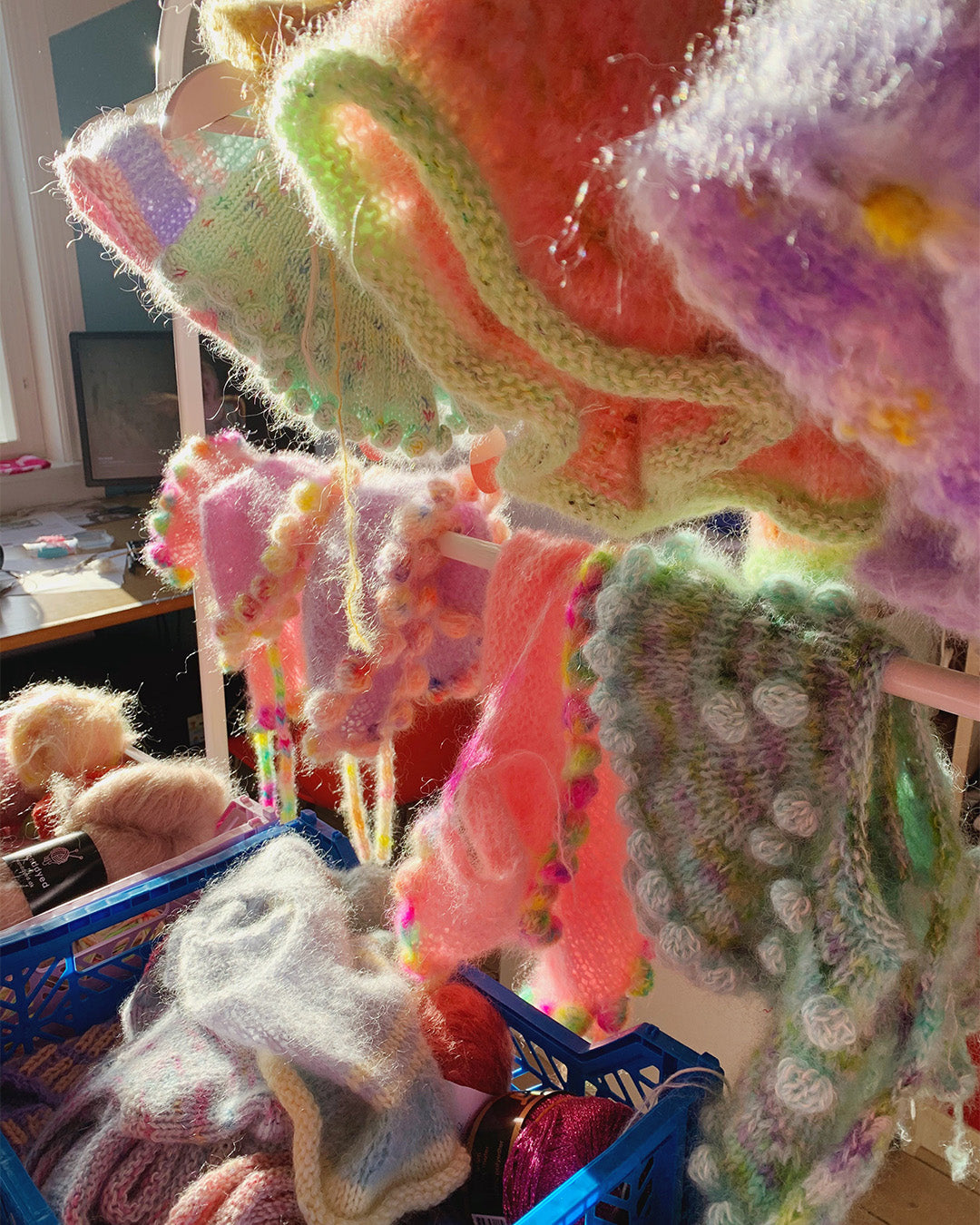 A close-up of fluffy, colorful knitwear and yarns in various stages of the crafting process, with sunlight filtering through, highlighting the textures and the lively chaos of a creative workspace.