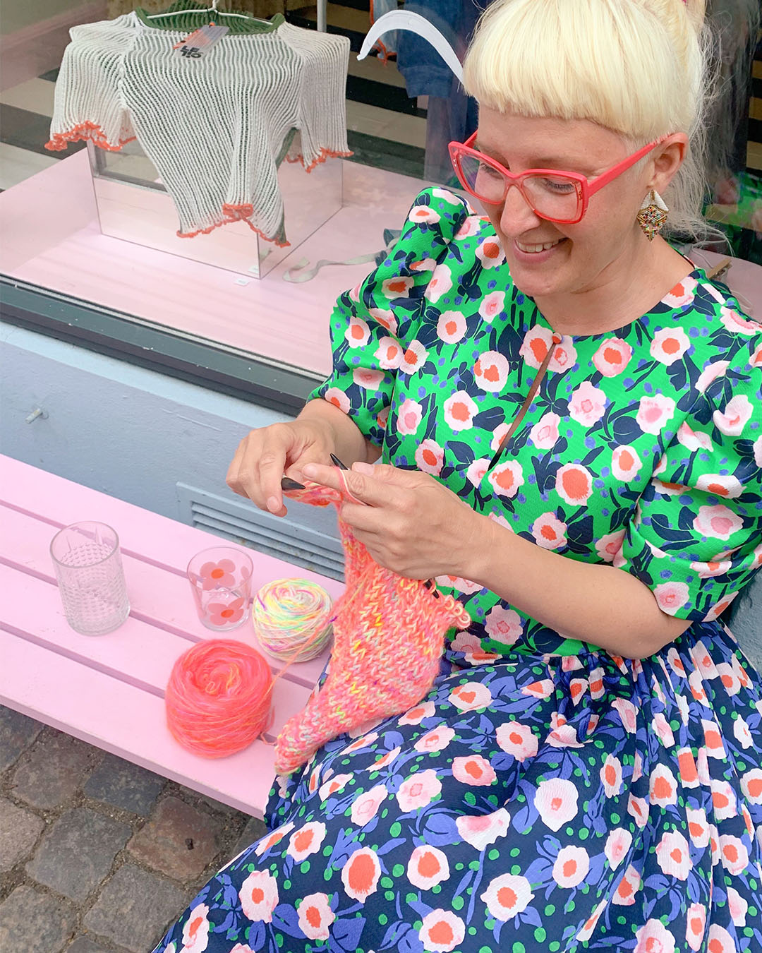 Sofie in a floral dress and bright red glasses sits outside, knitting with neon yarn, in front of a vibrant display window, her contented smile reflecting her passion for craft.