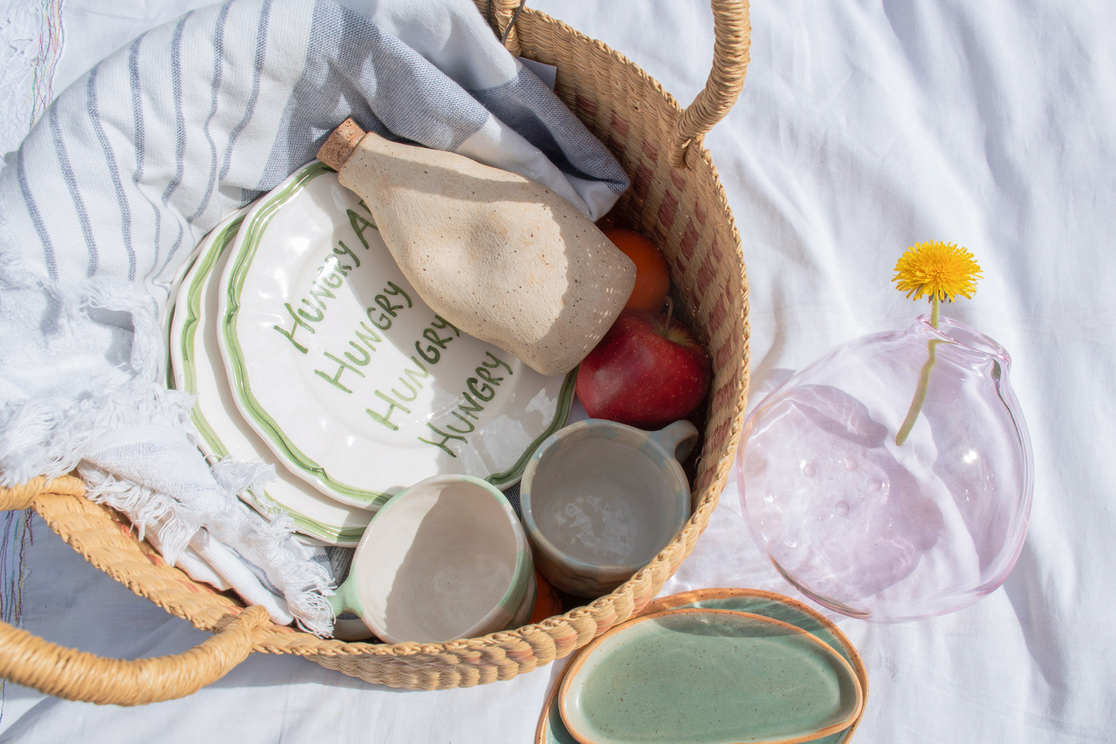 Inside a picnic basket, showcasing organic cotton napkins, a ceramic jug nestled among fresh fruit, with a glass carafe adorned with a single dandelion, emphasizing an environmentally-friendly outing.