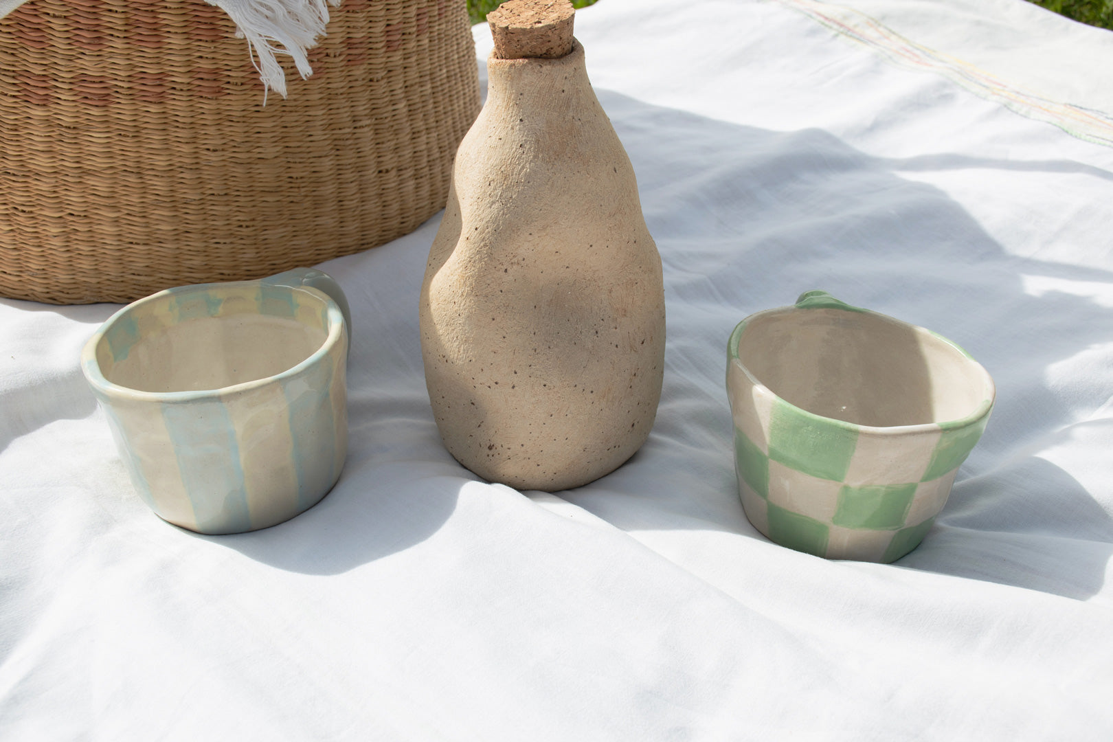 Handcrafted ceramic mugs in soft blue and green patterns alongside a natural clay jug, arranged on a white cloth amidst a woven basket.