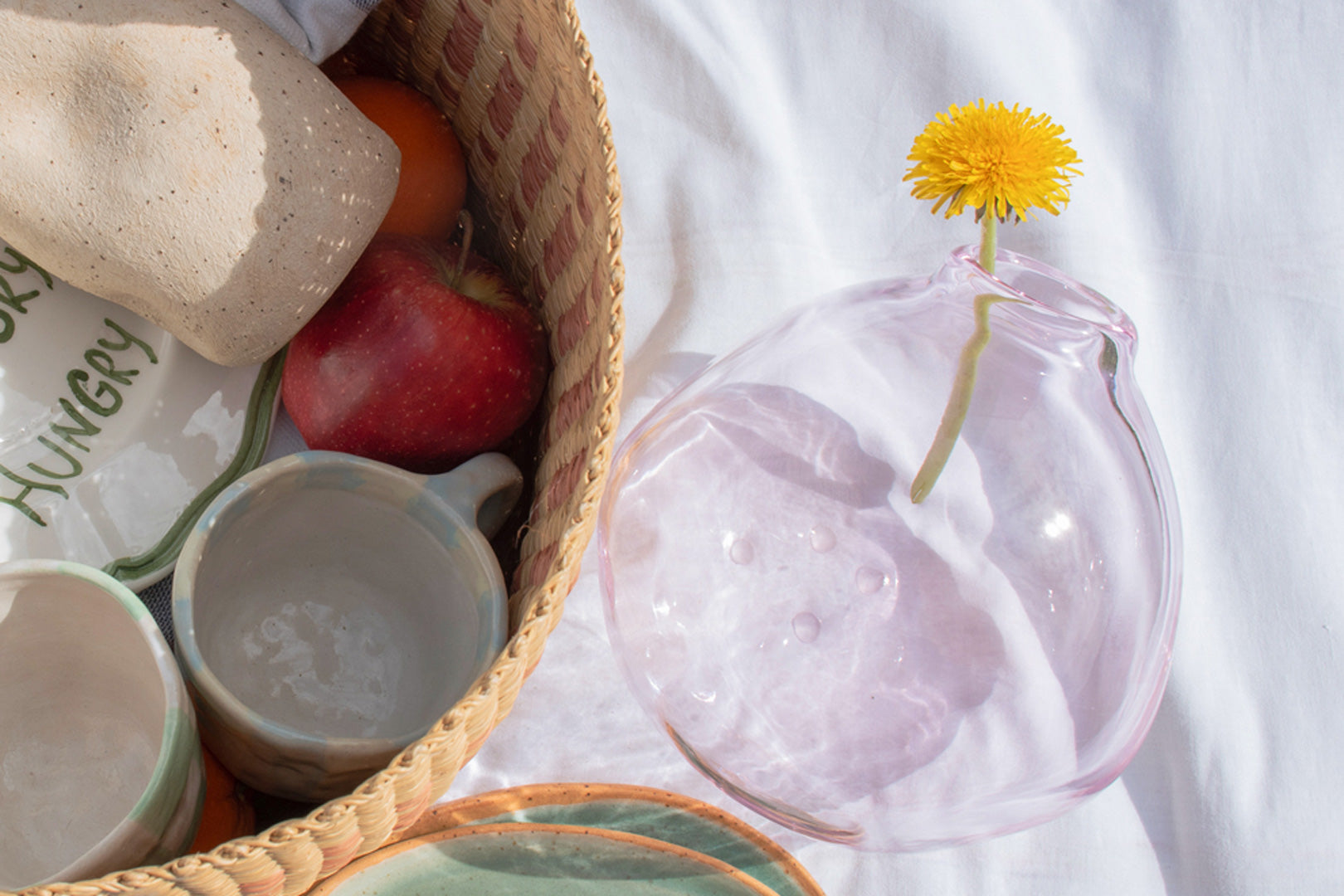 Inside a basket, aceramic jug, and cups nestle among fresh fruits, with a crystal pink glass carafe topped by a yellow dandelion.