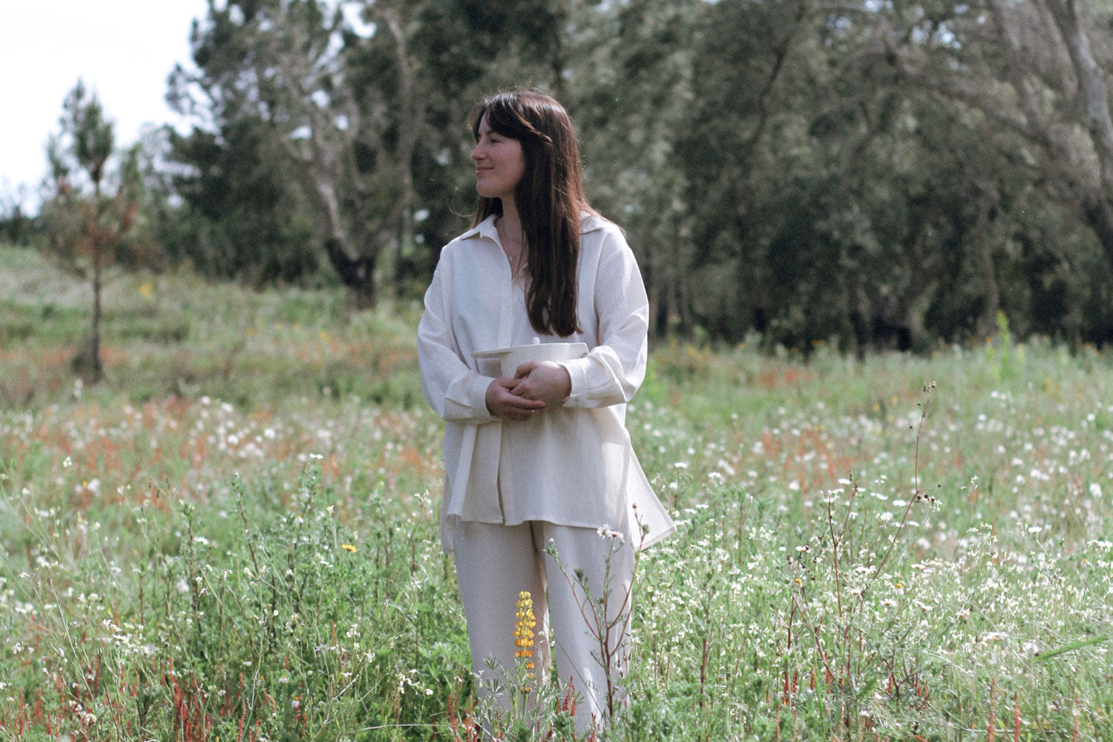 A woman in a white outfit stands serenely in a blooming meadow, holding a piece of handmade ceramic.