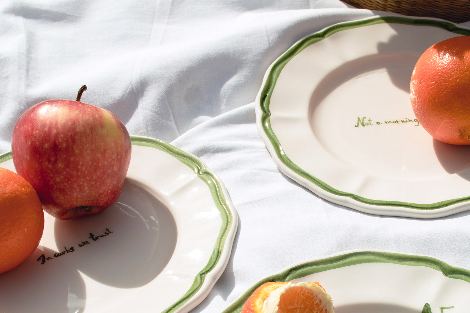 Picnic setting with fresh apples and citrus fruit on reusable ceramic plates with green borders, lying on a white blanket.