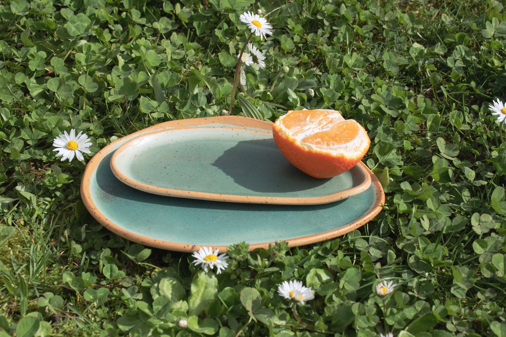 Ceramic plates in earthy teal hues, presented on a lush green clover field with a bright orange slice, showcasing eco-friendly outdoor dining options.