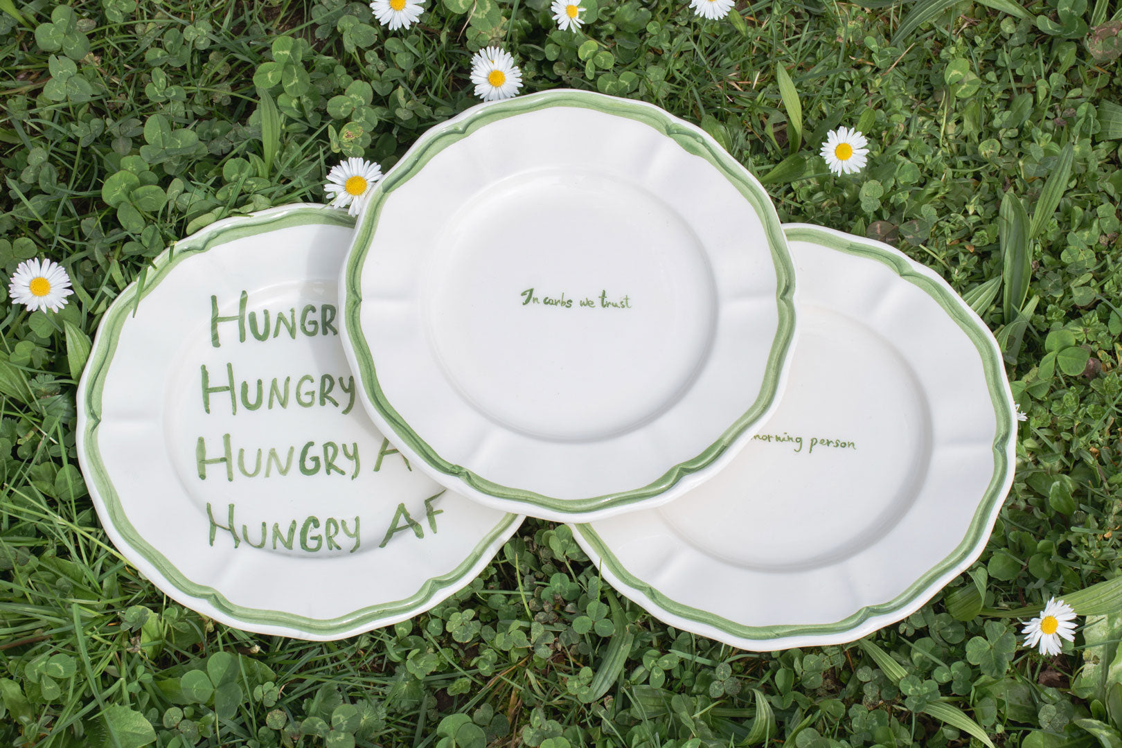 A series of white handpainted ceramic plates with green borders and whimsical food-related phrases, arranged on a bed of clover, displaying a playful approach to sustainable picnicware.