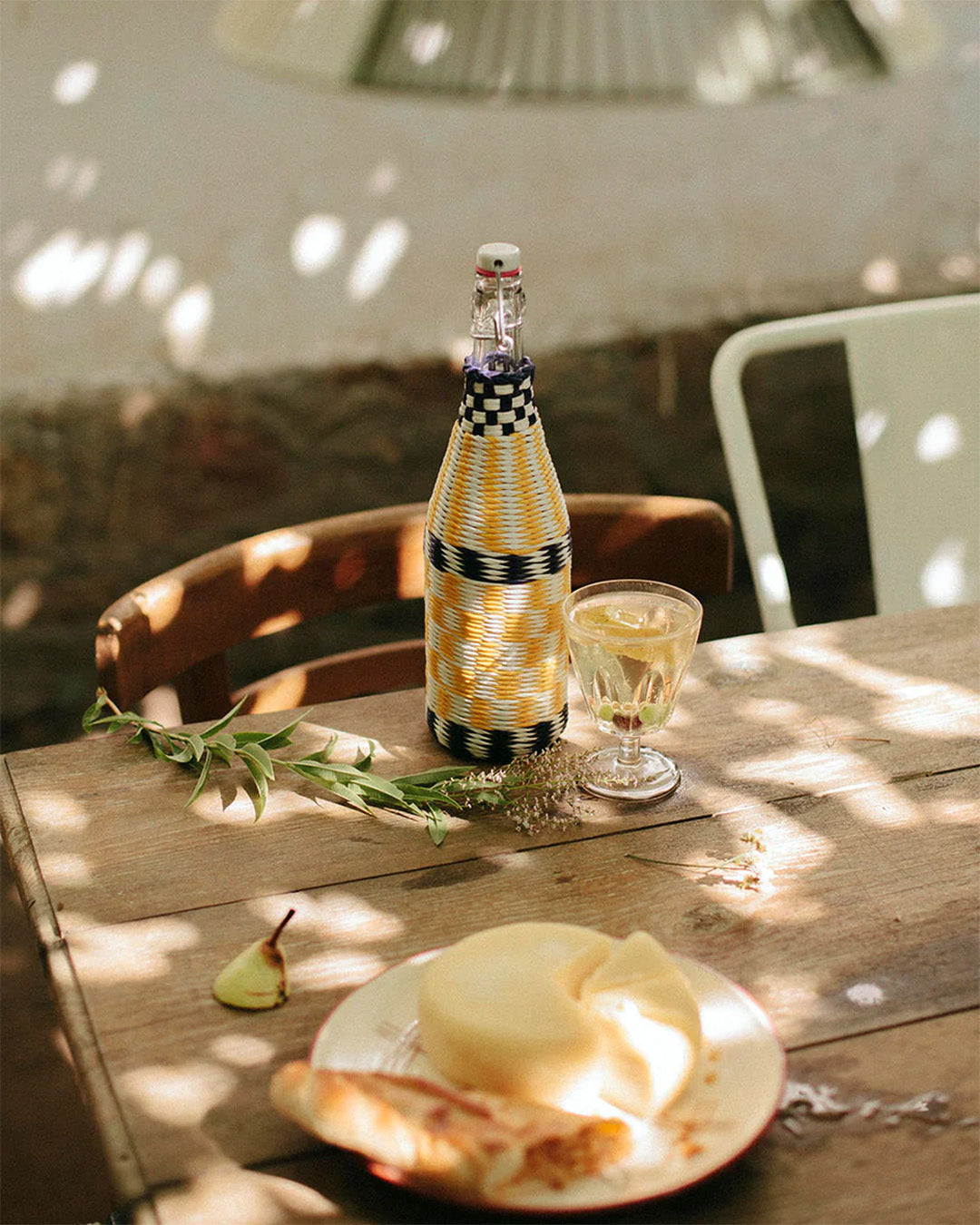 A bottle with handwoven recycled plastic wrap on a rustic wooden table accompanied by a simple glass and a plate of food.