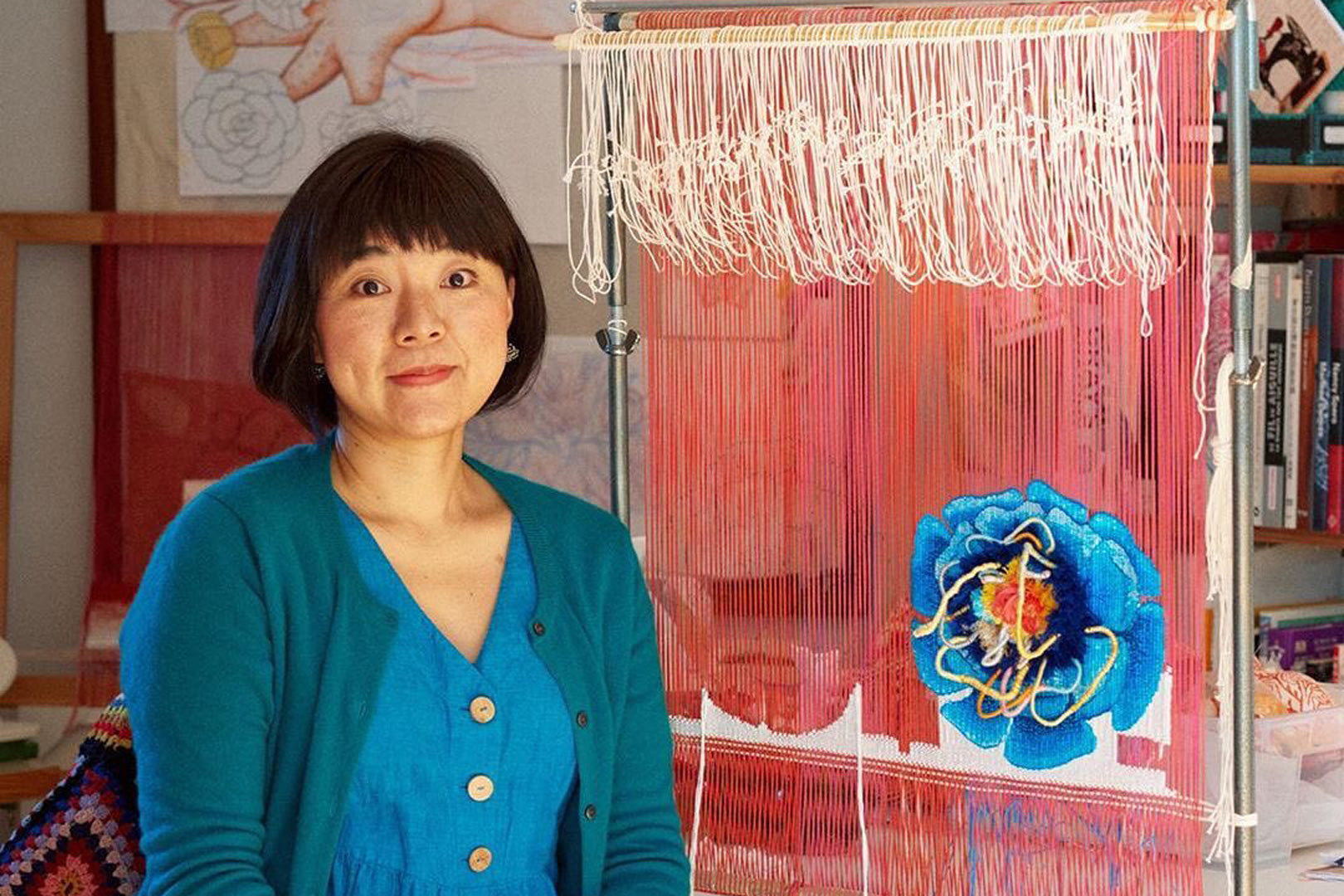 A female artist: Ema Shine,  in a turquoise cardigan smiling softly, sitting beside her colorful textile art on a loom in a studio filled with creative materials and art books.