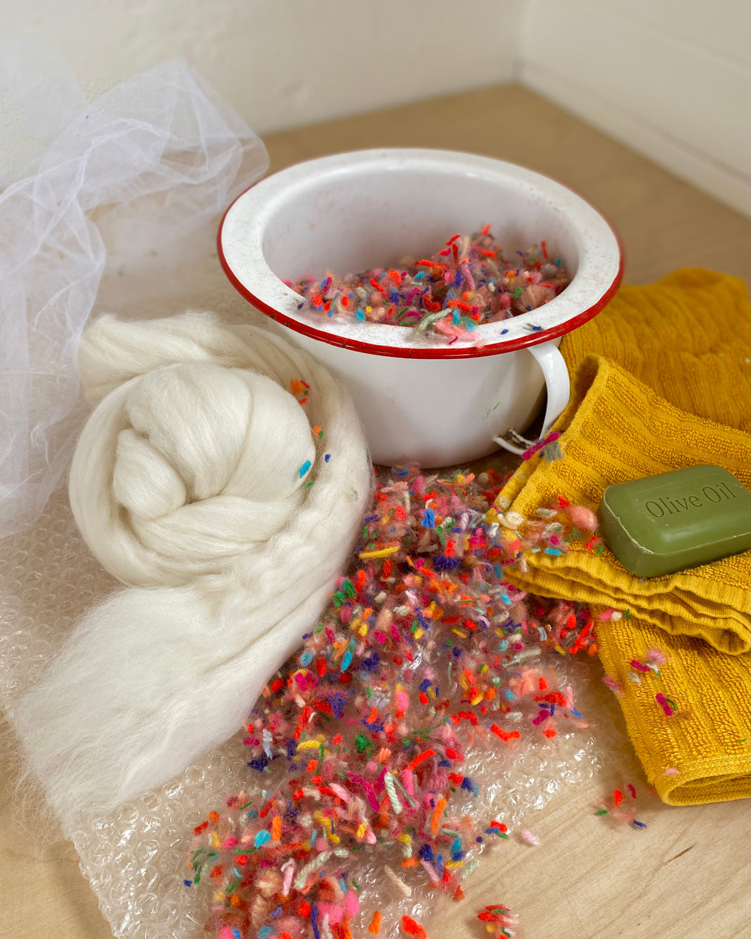 Bowl filled with colorful wet felting scraps alongside white wool and olive soap, demonstrating the felting materials.