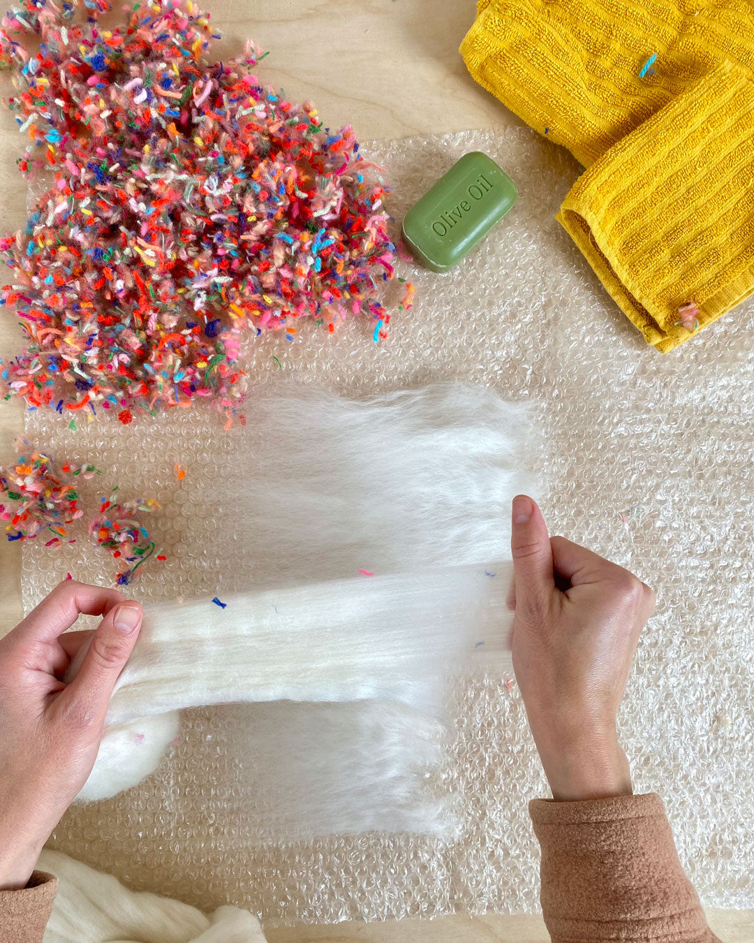 Craftsperson gently pulling at white wool fibres, preparing materials for a wet felting project on a textured surface.