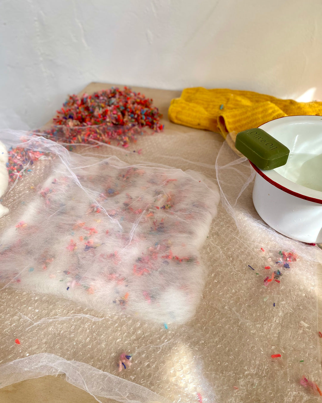 A snapshot of the wet felting process with multicolored scraps scattered over a sheet of white wool on a bubbled wrap.