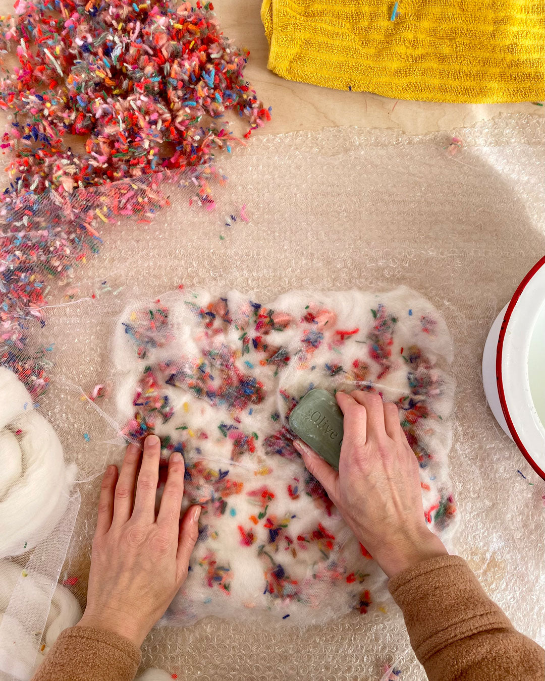 Crafting hands pressing a green olive soap bar onto wool roving amid a spray of colorful scraps during wet felting.