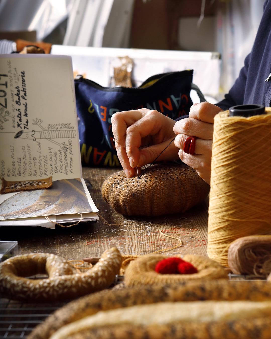 The process of Kate Jenkins sewing a textile piece of bread, with focused hands threading a needle, amid a spread of similar art pieces, capturing the essence of creative female craftsmanship.