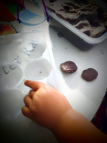 A toddler's hand presses people playdough into a plastic mold shaped like a leaf with already pressed playdough leaves on a white table.