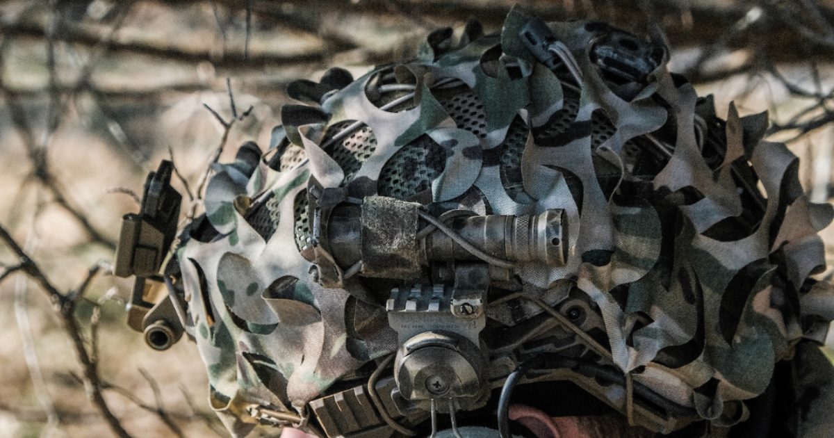 A solider hiding in the bushes wearing a camo green helmet scrim.