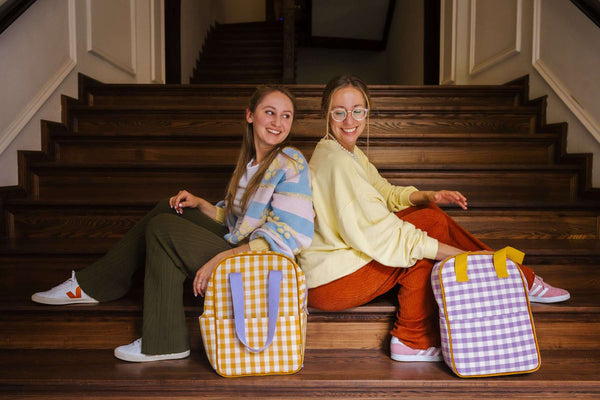 due amici seduti sulle scale e sorridenti. Davanti ci sono gli zaini a quadretti di Bettys Home