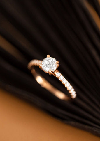 Round traditional diamond in a rose gold band with white diamonds, on a black fan and tan background