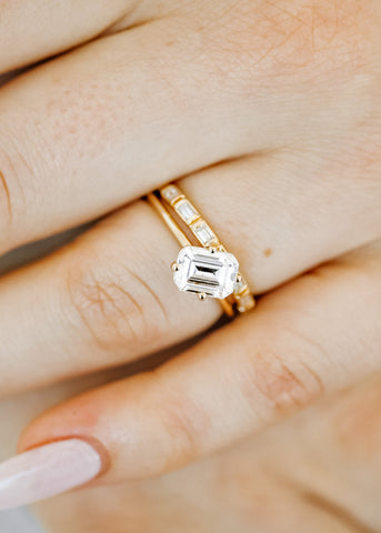 Rectangular clear moissanite set at a 45-degree angle on a plain yellow gold band, paired with a yellow gold wedding band with white, baguette-cut diamonds, on a fair-skinned hand