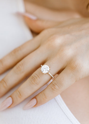 Round clear moissanite on a yellow gold band with white diamonds, on a fair-skinned hand with light pink nails