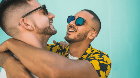 two men with arms around each other, embracing, with big smiles. blue background.