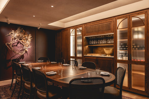 Image of a dark interior with brown walls. There is a large wooden dinning room table and dried flower walling hanging on the wall at the head of the table.