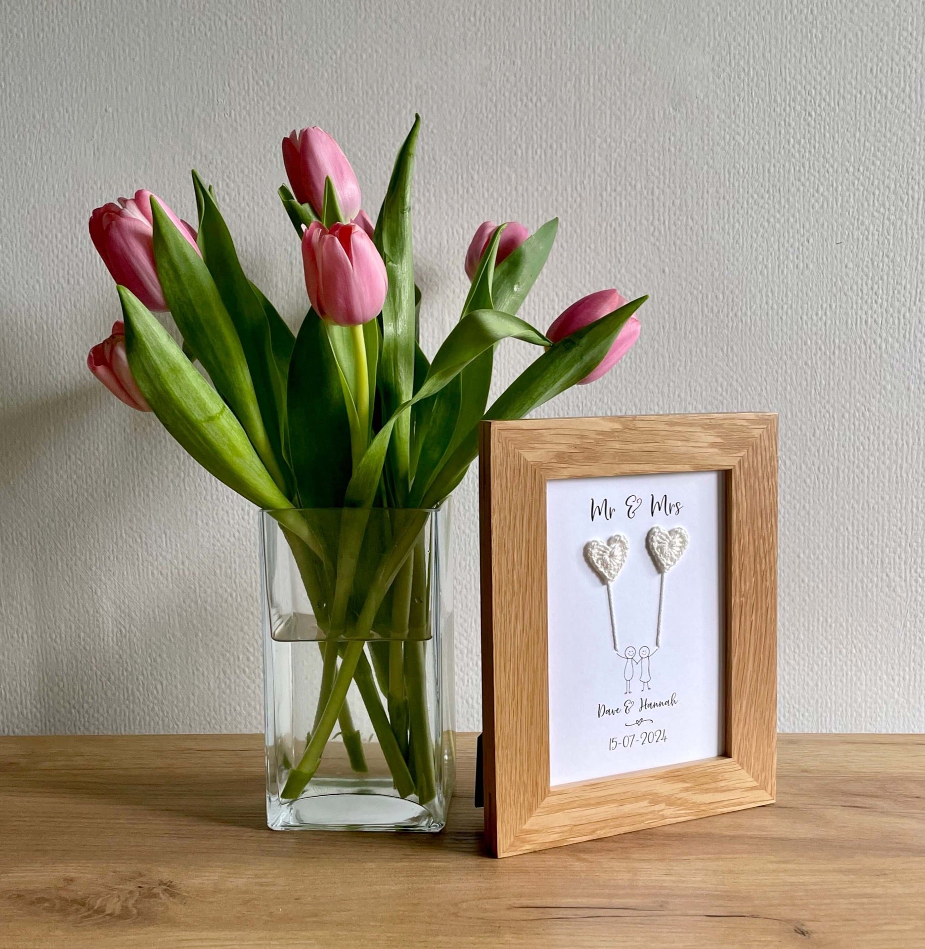 A personalised wedding card with ivory coloured crochet hearts framed in an oak frame. The frame sits next to a glass vase of pink tulips. 