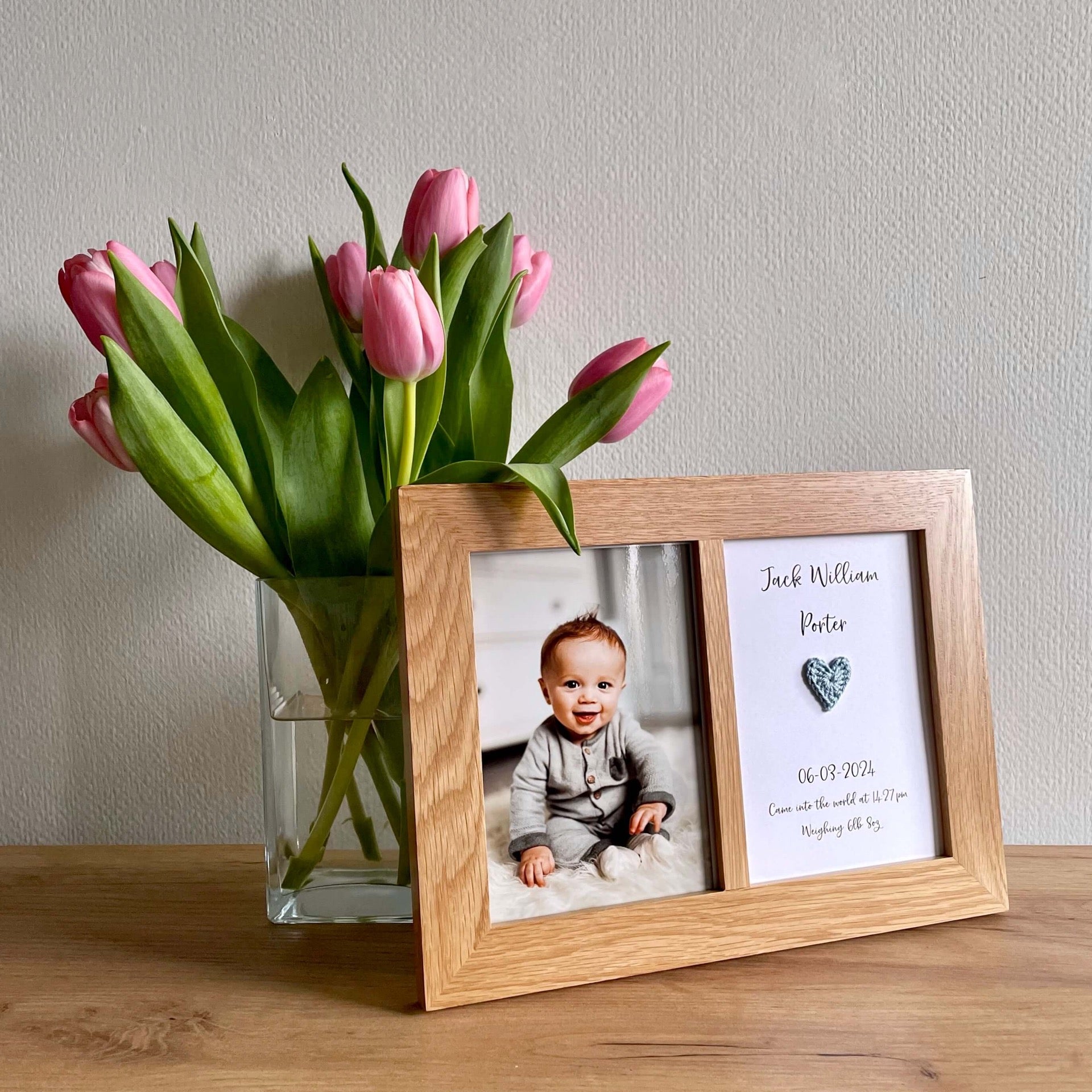 A personalised new baby boy card in an oak frame alongside a vase of pink tulips.