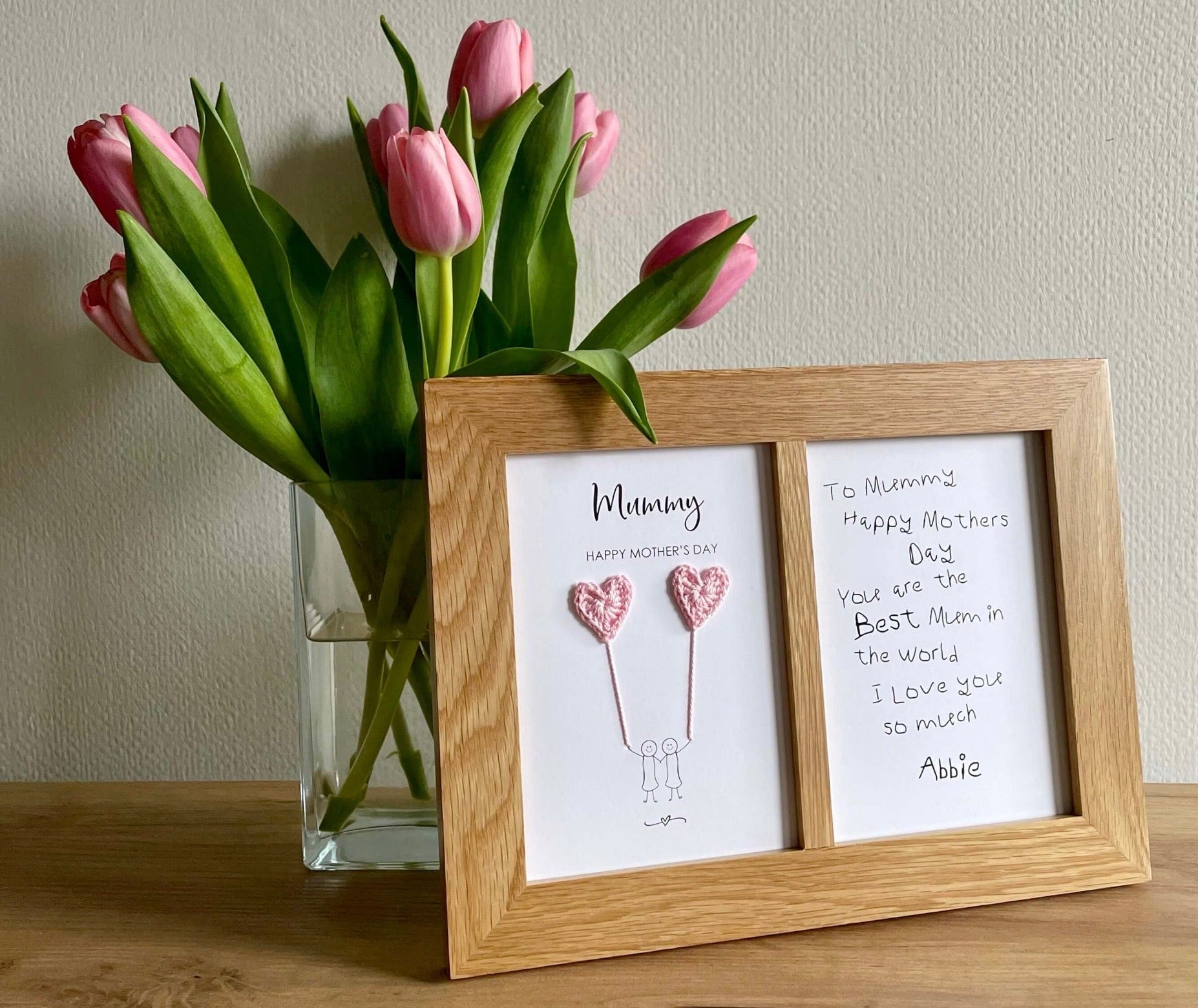 A Mothers day card featuring two pale pink crochet hearts has been framed in a double aperture oak frame. The front of the card is framed on the left and on the right is the heartfelt handwritten message from the child. The frame is placed next to a glass vase of pink tulips.