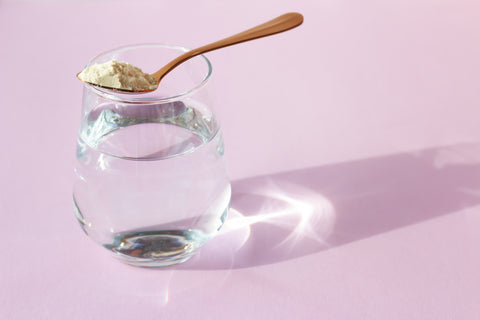 Collagen powder in a spoon and a glass of water on a pink background