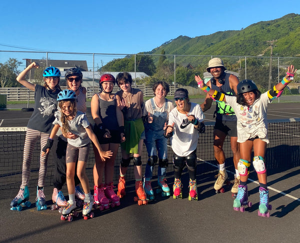 A group of children and adults on roller skates, smiling and cheering. Outside on a tennis court.