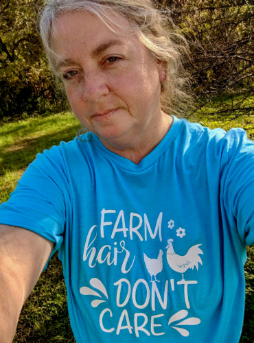 Cathy wears a teal t-shirt  with a chicken on it that reads Farm Hair Don't Care. 