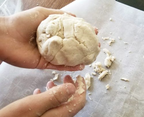 A pair of hands holding a dough ball. Crumbles of dough lay on a piece of parchment paper in the background.