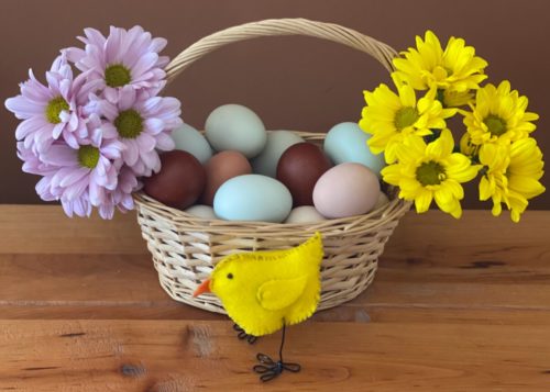 A DIY felt chicken with wire feet stands on a table in front of a woven basket filled with colorful eggs and fresh flowers.   