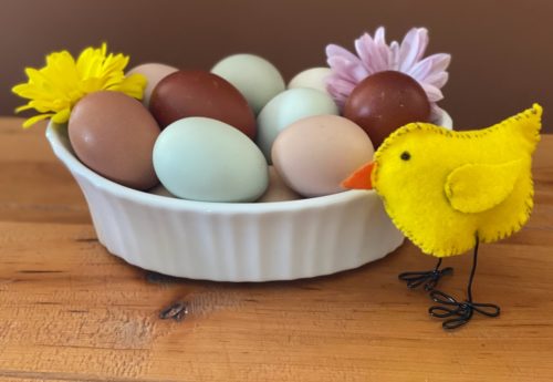 A DIY felt chicken with wire feet stands on a table in front of a white bowl filled with colorful eggs and fresh flowers.   
