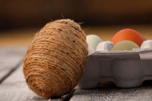 A jute bunny egg rests on an egg carton that is filled with colorful eggs. 