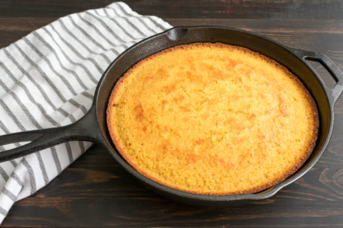 A skillet pan is laid on a wooden counter and has fresh cornbread in it. This cornbread makes an excellent cold-weather treat.