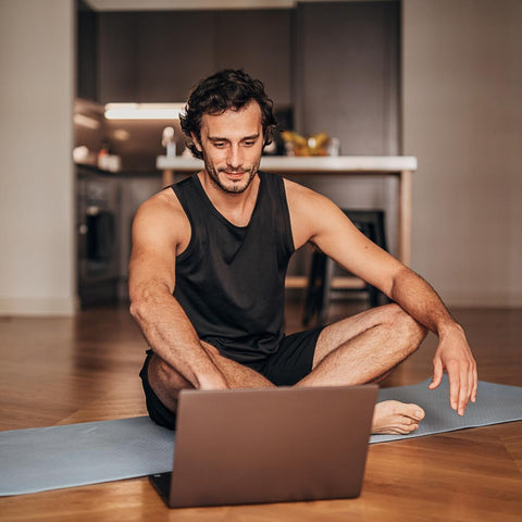 Muscular man sitting on yoga mat looking at laptop home workout