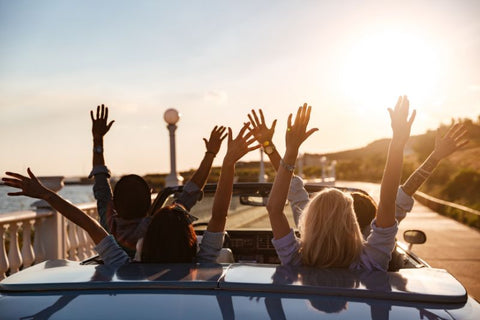 view from behind of open top car of people driving into sunset with hands in the air