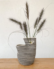 a black and white striped basket with two compartments is filled with furry grass stems and sits atop a wooden table with a white chipboard background