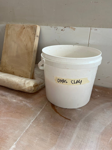 Image of bucket labelled "Oma's clay" sitting on wooden studio bench.