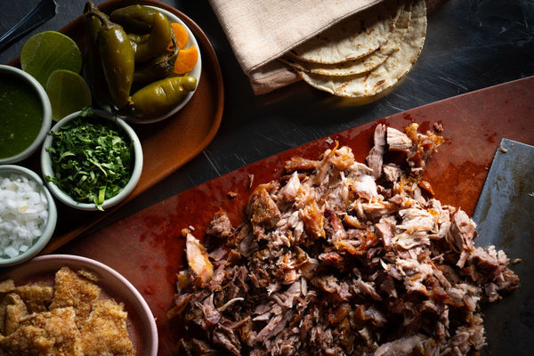 Cutting board with chopped carnitas, fresh tortillas, and garnishes like cilantro, onion, jalapeños on the side