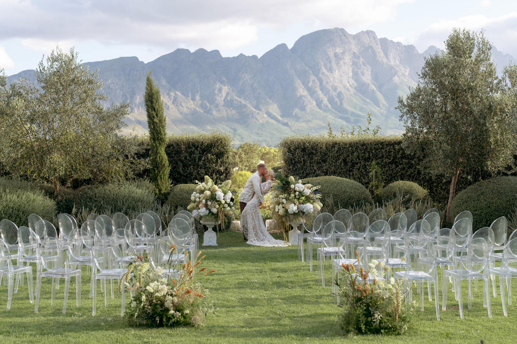 A couple kissing at the end of the aisle, surrounded by flowers and clear chairs. The ceremony is outside and there's mountains in the background.