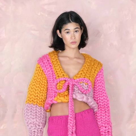 A model wearing the Rose Colossal Knit Cardigan from Hope Macaulay, which is knitted with pink, purple and orange yarn with a bow.